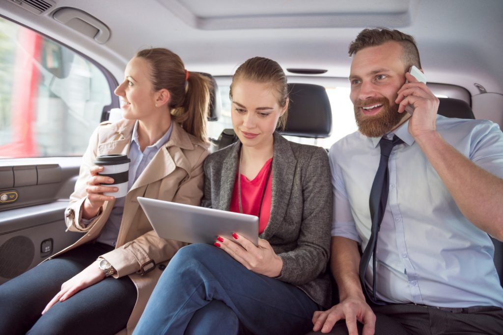Three professionals sit in the back of a car, one holding a coffee, another using a tablet, and the third talking on a smartphone.