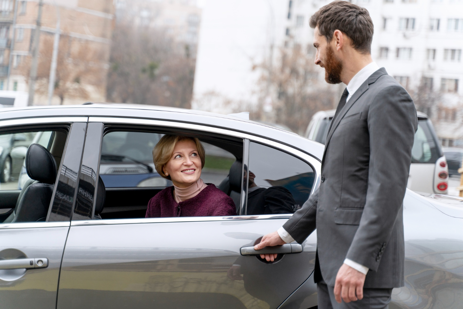 A man in a suit opens the car door for a woman inside, set against an urban backdrop.