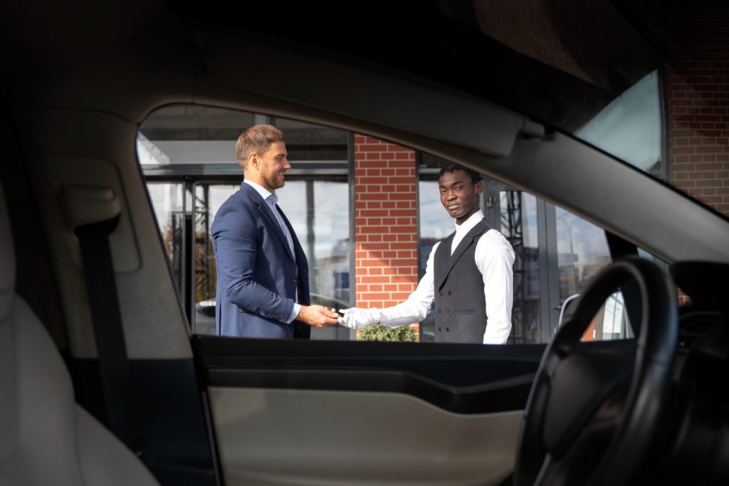 Two men in formal attire shake hands outside a modern building, viewed from inside a car.