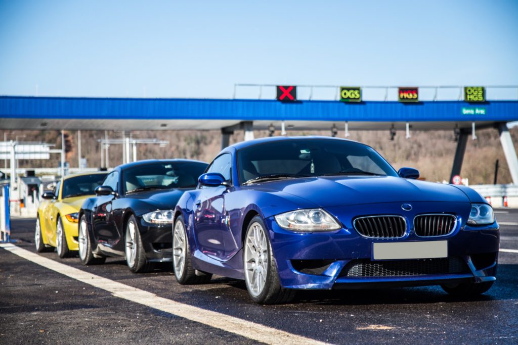 A lineup of four sports cars, including a blue BMW Z4, at a toll booth with electronic signage overhead and a clear blue sky.